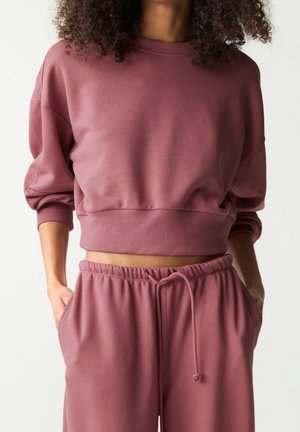 Femme portant un sweat-shirt court mauve et un pantalon de jogging assorti à cordon, mains dans les poches sur un fond uni.