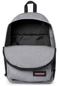 Mochila de tela gris con cierre de solapa, bolsillo delantero y un compartimento de malla en el interior. Presenta cremalleras negras y un logo en la parte frontal.