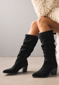 Bottes montantes en daim noir avec un design décontracté, bout pointu et talon carré. Détail de fermeture éclair latérale visible.