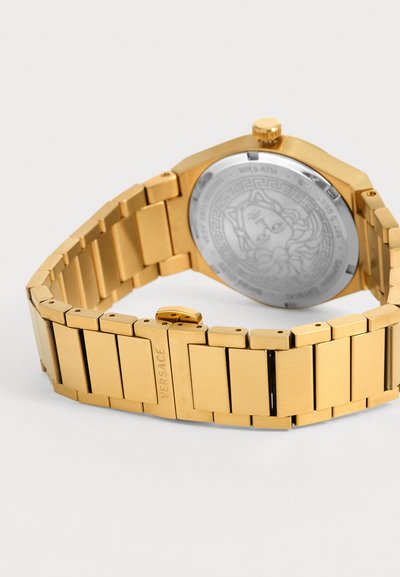 Bracelet de montre en métal ton or avec fermoir gravé Versace, montrant le fond en acier inoxydable avec le logo tête de Méduse.