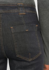 Jeans en denim foncé avec des coutures orange, dotés d'une poche arrière et d'une taille haute. Le tissu présente une texture lisse et une coupe classique.