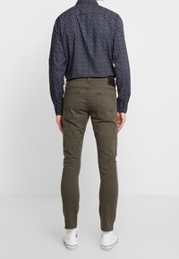 Persona con pantalones ajustados de color verde oliva, camisa de manga larga azul marino con estampado y zapatillas blancas altas, de pie de espaldas.