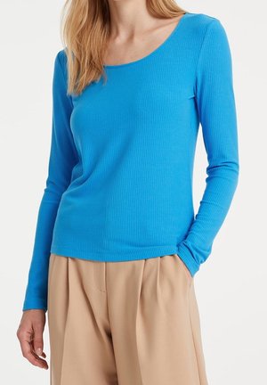Femme portant un haut bleu moulant à manches longues côtelé, rentré dans un pantalon beige ample, une main dans la poche, debout devant un fond blanc.