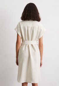 Abito in lino beige chiaro con maniche corte, taglio rilassato e vita con cintura. Il retro presenta un yoke e una leggera texture.