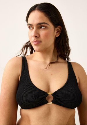 Kvinde med langt mørkt hår iført en tekstureret sort bikini-top med en rund frontlås, der ser mod venstre mod en neutral baggrund.