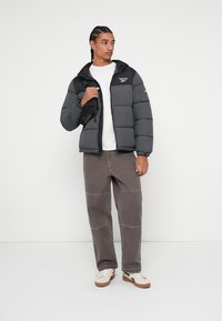 Grijze pufferjack met een capuchon, voorzien van een ritsluiting en een logo op de borst. Gepaard met bruine wijde broek en witte sneakers.