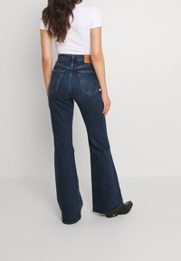 Nainen, jolla on yllä tummansiniset korkeavyötäröiset leveälahkeiset farkut ja valkoinen lyhythihainen paita, seisoo yksinkertaista vaaleataustaa vasten.