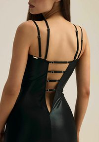 Czarna sukienka bodycon z cienkimi, regulowanymi ramiączkami, głębokim dekoltem na plecach z metalowymi akcentami oraz gładką, błyszczącą teksturą.