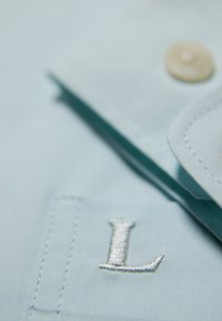 Camicia azzurra chiara con una "L" ricamata sul taschino, colletto arrotondato e un bottone crema sulla cucitura della spalla. Tessuto liscio.