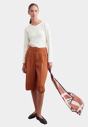 Mujer con una camiseta blanca de manga larga y pantalones cortos marrones hasta la rodilla, sosteniendo una bufanda marrón y blanca con patrón, de pie frente a un fondo liso.