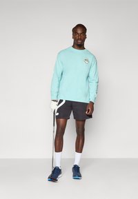Long-sleeve mint shirt with a small graphic on the chest, paired with black shorts featuring a white abstract design. Golf club in hand.