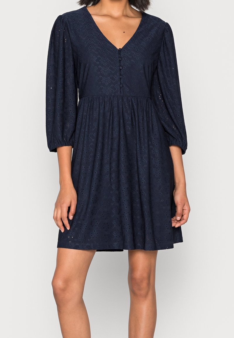 Robe bleu marine avec un col en V, des manches ballons et un bouton devant. Fabriquée en tissu texturé avec un motif à œillets et une jupe évasée.