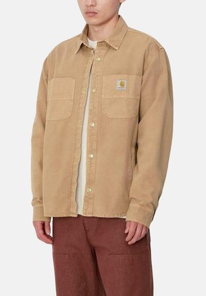 Chaqueta de camisa de pana color beige con cierre de botones en el frente, dos bolsillos en el pecho y un parche de la marca. Textura suave, corte relajado y mangas largas.