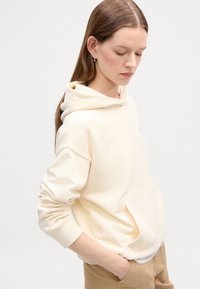 Haut à capuche de couleur crème avec une poche avant et un cordon de serrage. Tissu doux, coupe décontractée et poignets côtelés. Porté avec un pantalon beige.