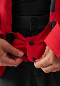 Giacca rossa con accenti neri, caratterizzata da una texture liscia, grandi bottoni e dettagli in rete attorno alle spalle, con chiusura frontale.