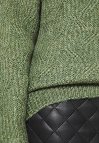 Pull en tricot vert avec un motif texturé, présentant des poignets côtelés. En dessous, un article en cuir matelassé noir est visible.
