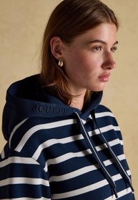 Young woman with brown hair wearing gold drop earrings and a navy blue and white striped hooded sweatshirt with "JOULES" on the hood.