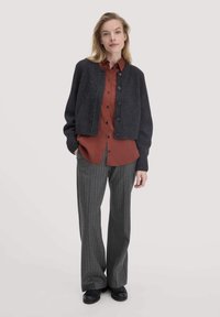 Dunkelgrau geschnittene Cardigan aus Wollmischung, texturiert gestrickt, mit Knopfleiste. Darunter ein rostfarbener Hemd mit Knöpfen. Gestreifte graue Stoffhose.