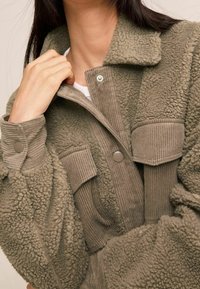 Vachte olijfgroene jas met een kraag, drukknoopsluitingen en geribbelde zakaccenten. Heeft een gestructureerde stof voor extra warmte.