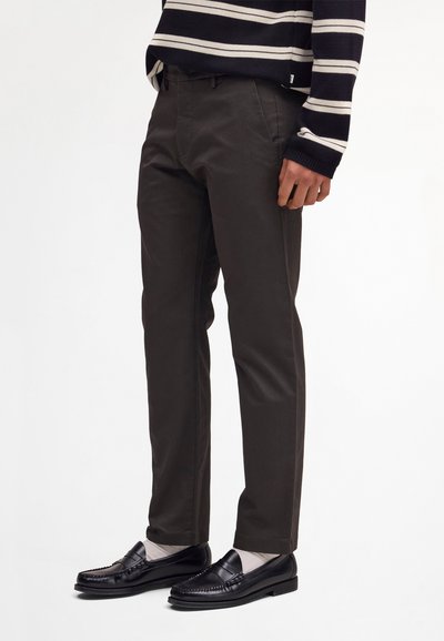 Homme portant un pantalon droit foncé, des mocassins noirs, des chaussettes blanches et un pull rayé noir et blanc, debout devant un fond uni.