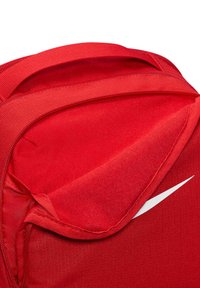 Czerwony plecak Nike z miękką fakturą, zaokrąglonym wierzchem i białym logo swoosh z boku. Zamek błyskawiczny pokazany częściowo otwarty.