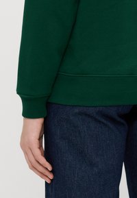 Grønn genser med ribbestrikket hals og ermer, kombinert med mørkeblå denimjeans. Stoffet ser mykt ut med en jevn tekstur.