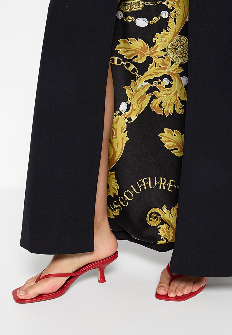 Personne portant un pantalon large noir révélant un vêtement intérieur à motifs noirs et dorés et des sandales à talons kitten rouges avec des lanières.