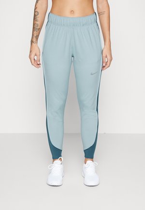 Personne portant un pantalon de sport Nike bleu clair et vert sarcelle foncé ainsi que des baskets blanches, debout avec les bras détendus le long du corps.