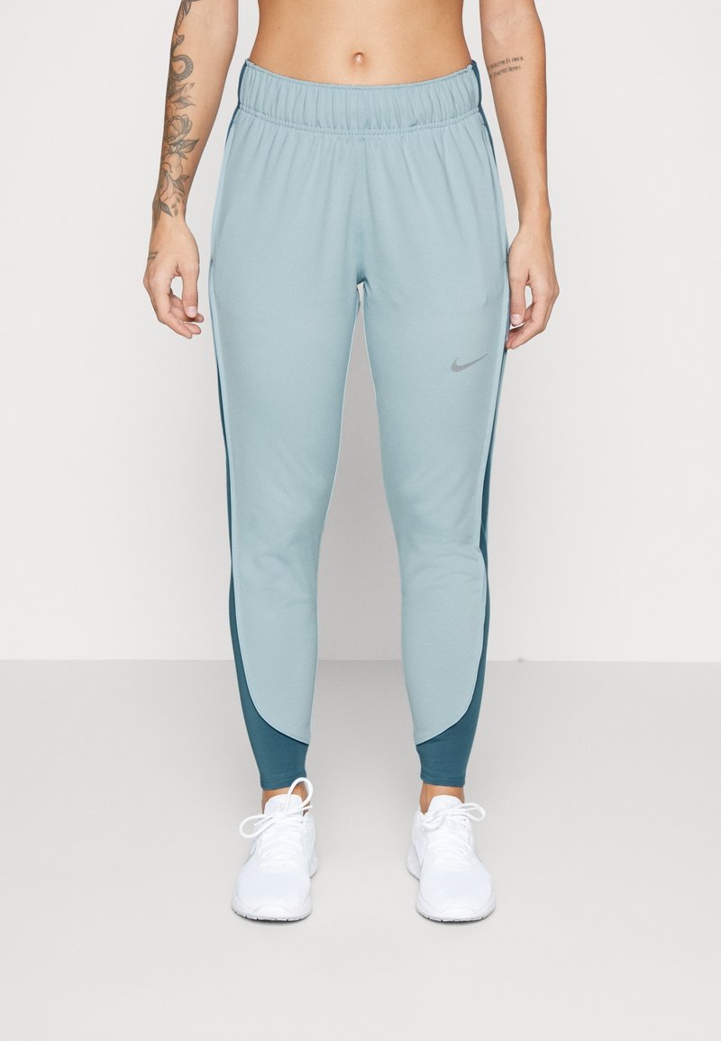 Personne portant un pantalon de sport Nike bleu clair et vert sarcelle foncé ainsi que des baskets blanches, debout avec les bras détendus le long du corps.