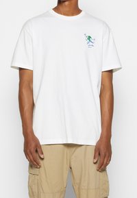 Mies joka pukeutuu valkoiseen lyhythihaiseen T-paitaan, jossa on pieni vihreä ja sininen pilakammo Maapallo skeittaamassa rinnassa, yhdistettynä beigeihin taskushortsseihin.