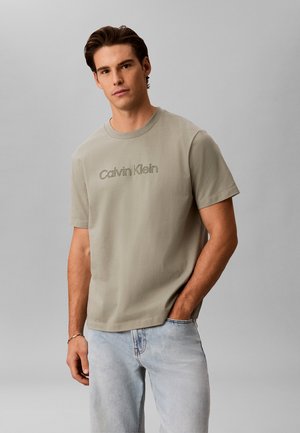 Mann mit braunen Haaren, der ein hellbeiges Calvin Klein T-Shirt und hellblaue Jeans trägt, steht mit einer Hand in der Tasche.
