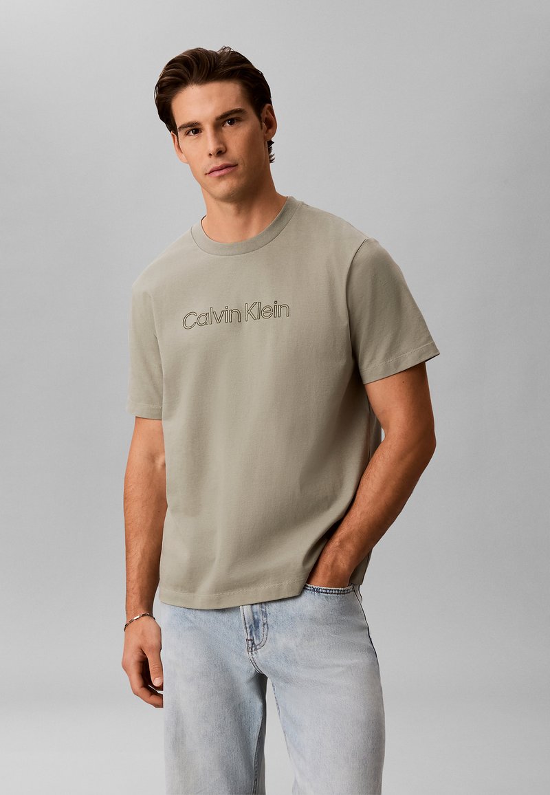 Homme aux cheveux bruns portant un t-shirt Calvin Klein beige clair et un jean bleu clair, debout avec une main dans la poche.
