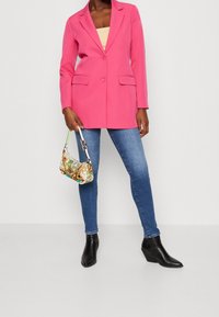 Veste rose vif ajustée avec un bouton unique, deux poches avant et une texture lisse. Associée à un jean bleu et un sac à main à motif.