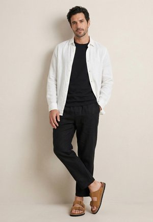 Homme debout contre un mur beige, portant une chemise blanche ouverte sur un t-shirt noir, un pantalon noir et des sandales marron à double bride.