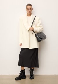 Cappotto di lana bianco con colletto sciallato, maglione beige, gonna di raso nera e stivali neri con lacci. La donna tiene una borsa a tracolla in pelle nera.