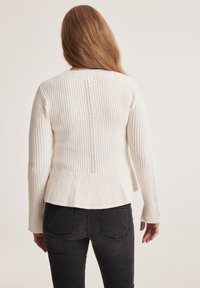 White knitted sweater with ribbed texture, featuring a small lace panel down the center back and long sleeves. Paired with black jeans.