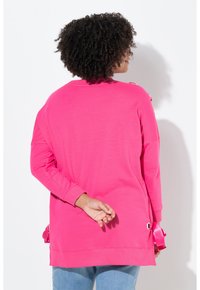 Oversized roze sweatshirt met lange mouwen en afgeronde zoom, voorzien van decoratieve lintaccenten en metalen ringen aan de zijkanten.