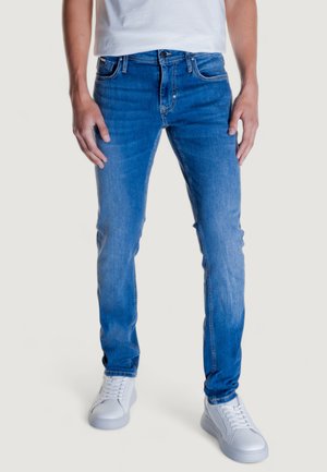 Pantalones de mezclilla azul con un corte slim, que presentan bolsillos frontales, un cierre de botón y detalles de desgaste sutiles. Combinados con zapatillas blancas.