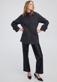 Cappotto in misto lana grigio scuro con colletto oversize, tasche anteriori e polsini abbottonati, abbinato a pantaloni in pelle nera e tacchi a punta.
