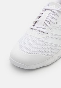 Białe sportowe buty z teksturowaną siateczkową cholewką, gładkim gumowym noskiem i płaskimi sznurowadłami z akcentami logo. Trwała konstrukcja do aktywnego użytkowania.