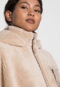 Veste en polaire beige et duveteuse avec un col haut, fermeture éclair à l'avant et poches latérales. Texture lisse avec une apparence douce et chaleureuse.