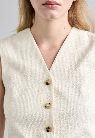 Haut sans manches en blanc cassé, texturé, avec un décolleté en V et trois grands boutons contrastants dans des nuances de crème et de noir.