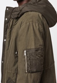 Olivgrön jacka med avtagbar huva, med en ficka med dragkedja på ärmen och tryckknappslås längs framsidan.