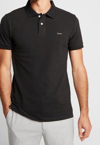 Polo noir en coton avec une finition texturée, doté d'un col, de deux boutons et d'un petit logo sur le côté gauche de la poitrine.