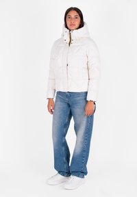 Witte gewatteerde pufferjack met een hoge kraag, rits aan de voorkant en twee zakken, gedragen met blauwe recht-toe-rechtaan jeans en witte sneakers.