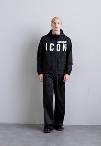 Schwarze Windbreaker-Jacke mit großem "ICON"-Text auf der Brust, kombiniert mit lockeren schwarzen Hosen. Geschmeidiger Stoff und entspannter Schnitt insgesamt.