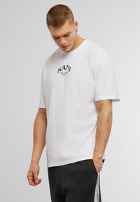 White cotton t-shirt with short sleeves and a crew neck. Features black "KANI Brooklyn NYC" text logo on the front. Casual fit.