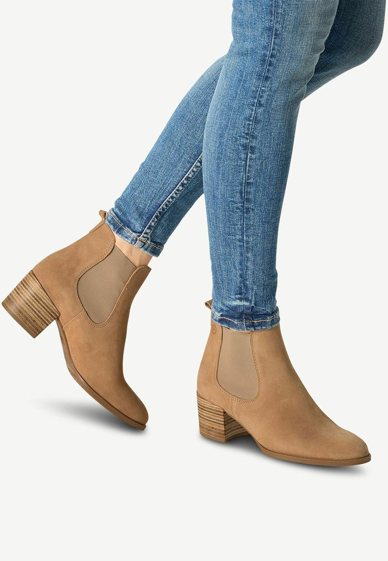 Botas de tobillo de ante color beige con paneles elásticos en los laterales, tacones de madera apilada y punta redondeada. Combinadas con jeans ajustados azules.
