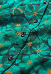 Trumpas rankovės marškiniai žaliame audinyje su gėlių raštu, kuriame yra dideli teal spalvos lapai ir maži oranžiniai žiedai, puošti juodais mygtukais.