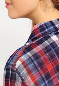 Chemise à carreaux rouge et bleu marine avec un col boutonné et un tissu doux et texturé. Présente un seul bouton sur la couture du col.